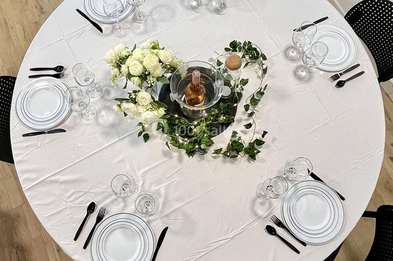Table ronde dressée avec une nappe blanche, vaisselle élégante, couverts noirs, fleurs blanches et bouteille au centre.