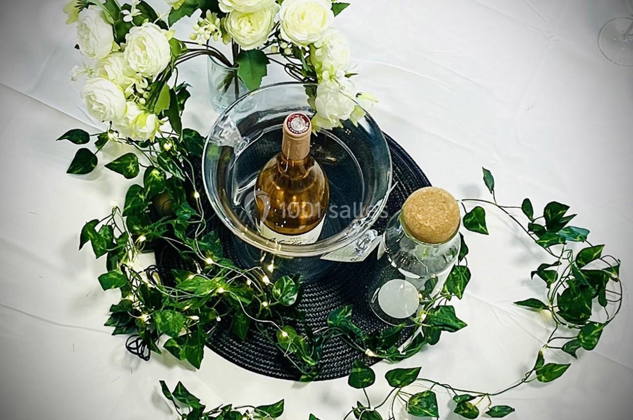 Centre de table avec une bouteille de vin rosé dans un seau, fleurs blanches, feuillage et guirlande lumineuse.