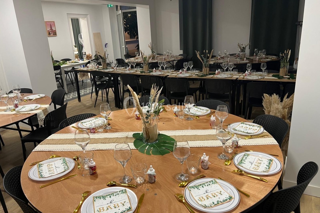Salle décorée pour un événement, avec des tables rondes dressées, ornées de centres de table végétaux et de vaisselle…