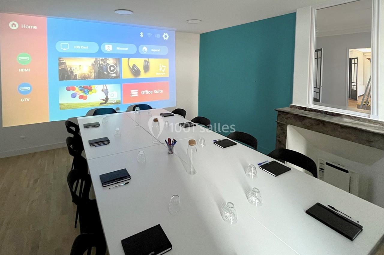 Salle de réunion moderne avec une grande table blanche, des carnets, un écran de projection allumé et un mur bleu.