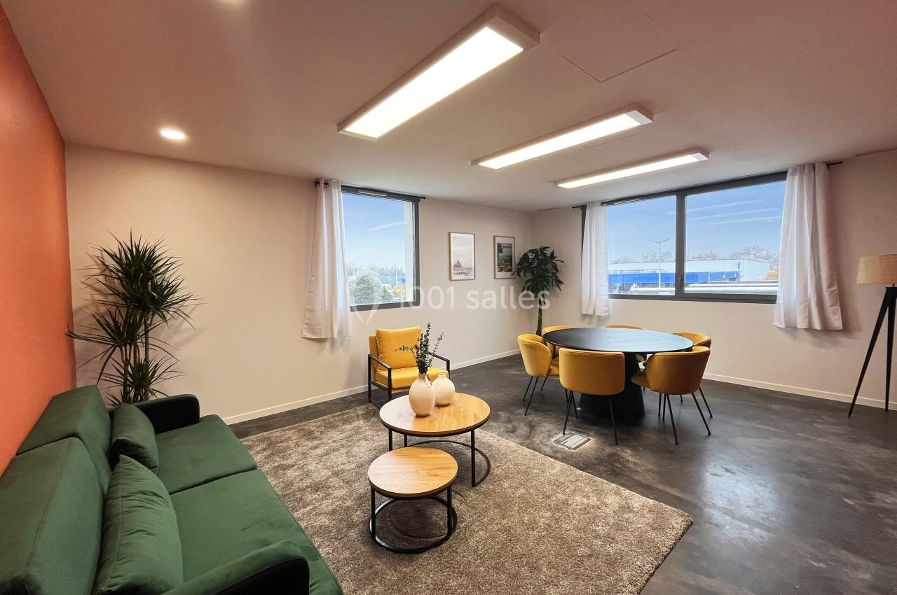 Salle lumineuse avec canapé vert, table basse, chaises jaunes autour d'une table, plantes et grandes fenêtres.
