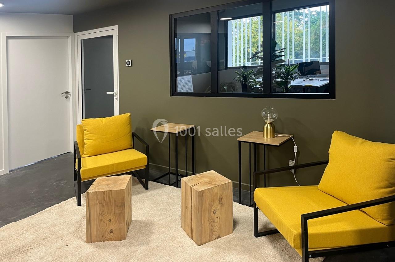 Espace d'attente avec deux fauteuils jaunes, tables en bois, lampe et fenêtre donnant sur un bureau.