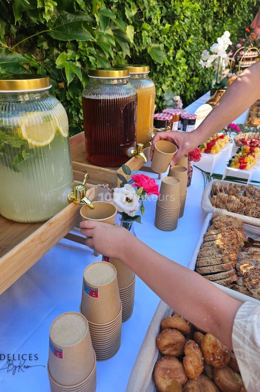 Distributeur de boissons avec citronnade, thé glacé et jus, entouré de viennoiseries et fruits sur une table en extérieur.