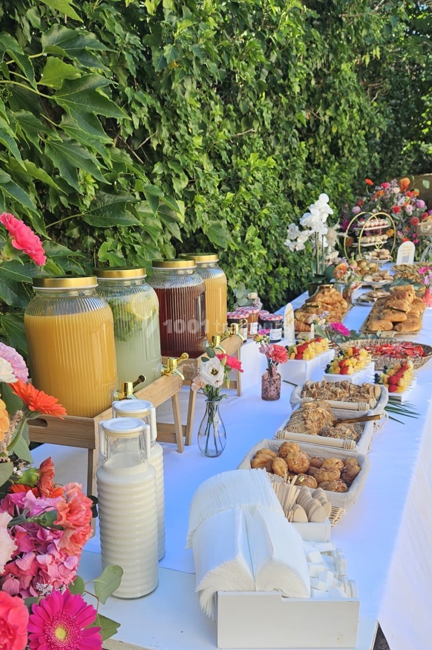 Buffet en extérieur avec boissons, viennoiseries, fruits et fleurs colorées sur une table blanche près d'un mur végétal.