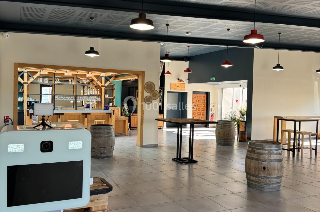 Salle lumineuse avec bar en bois, tonneaux décoratifs, tables hautes et borne interactive au premier plan.