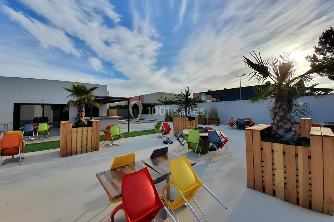 Terrasse extérieure avec tables en bois, chaises colorées et palmiers sous un ciel partiellement nuageux.