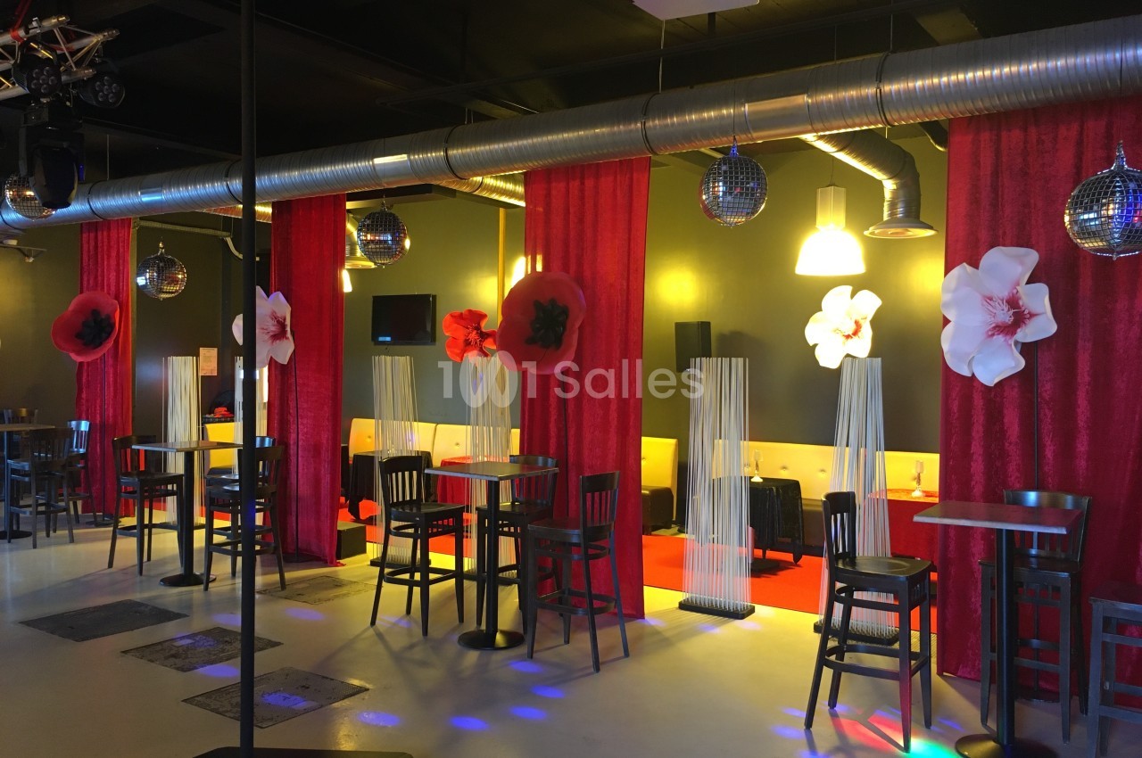 Salle décorée avec des tables hautes, rideaux rouges, fleurs murales et boules à facettes suspendues.