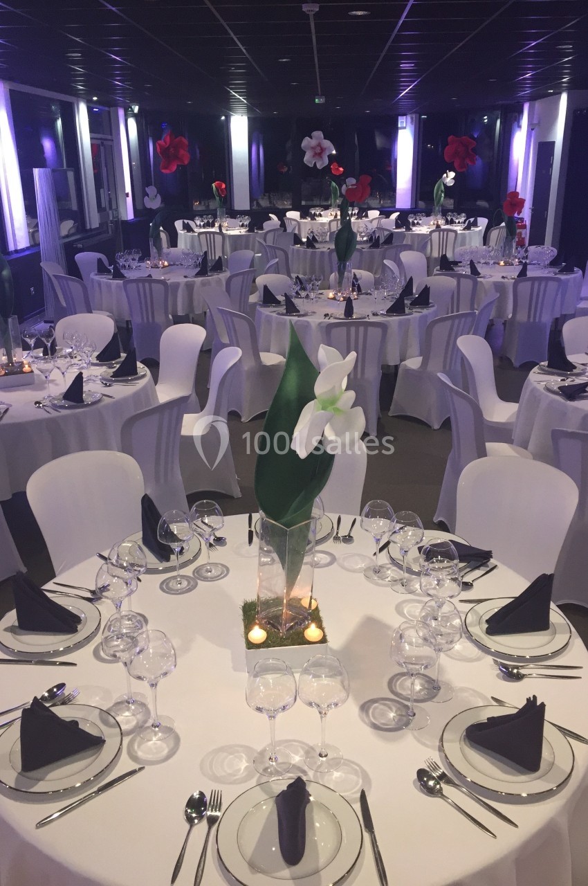 Salle de réception décorée avec des tables rondes, nappes blanches, serviettes noires et centres de table floraux.