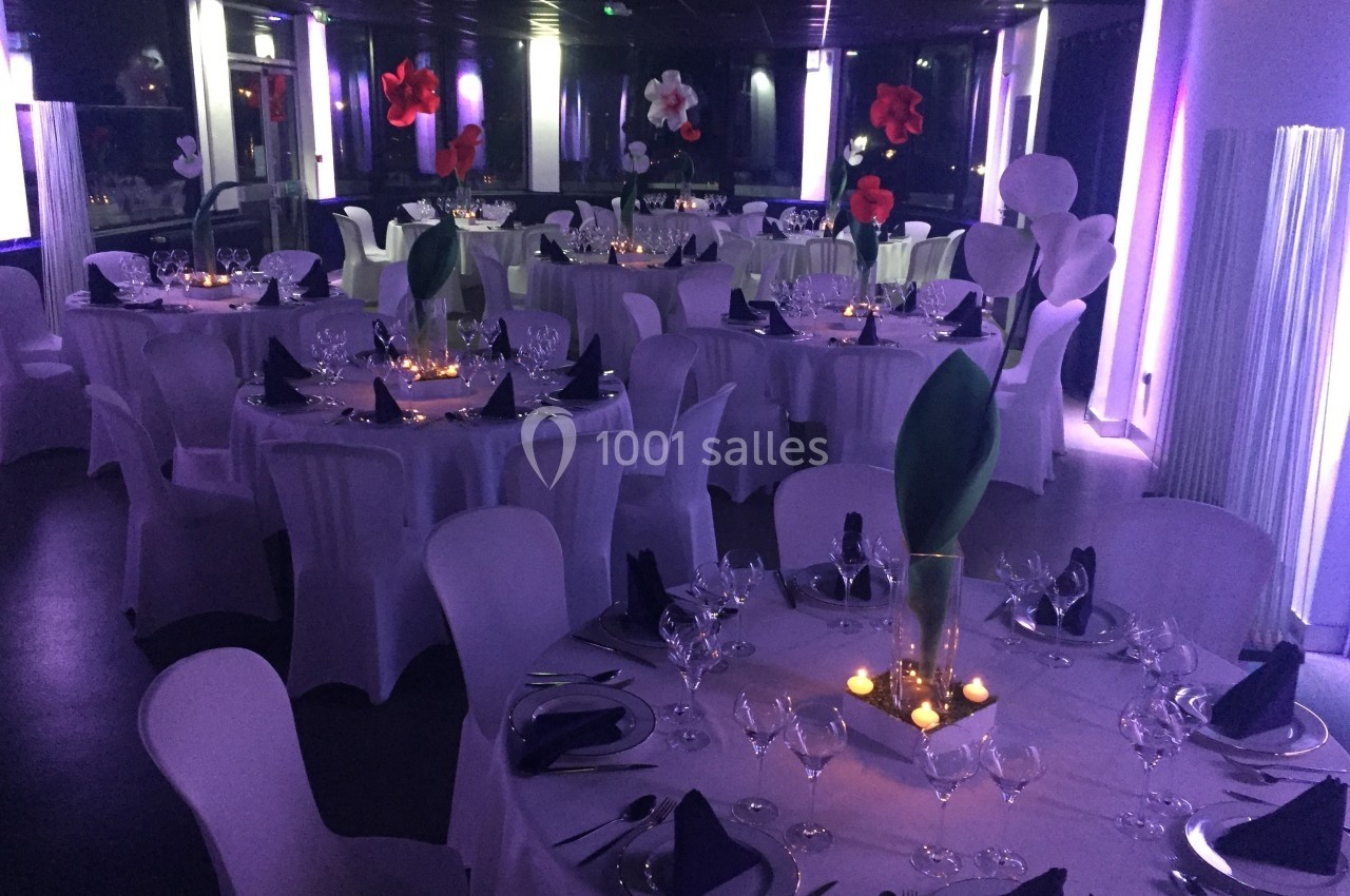 Salle de réception décorée avec des tables rondes, nappes blanches, serviettes pliées et éclairage tamisé violet.