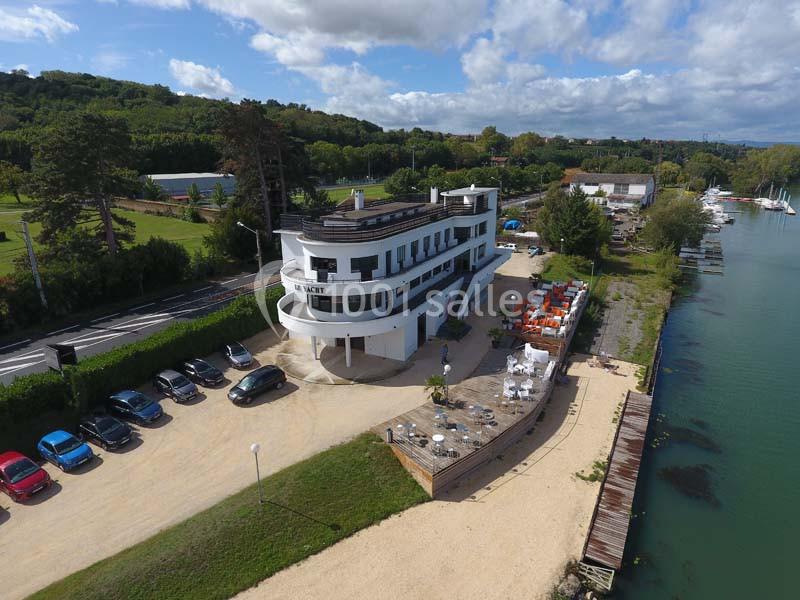 Vue aérienne d'un bâtiment moderne près d'une rivière, entouré de voitures, de terrasses et de verdure.