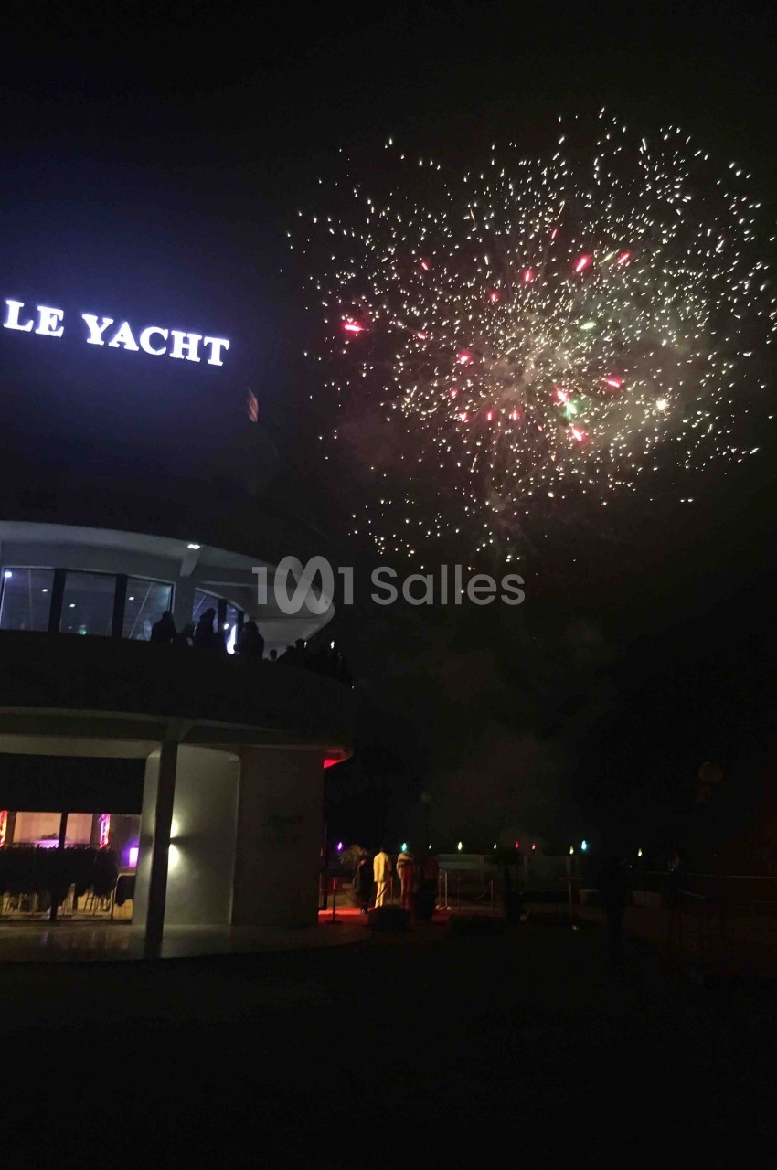 Feu d'artifice illuminant le ciel nocturne près d'un bâtiment éclairé avec l'inscription ’Le Yacht’.