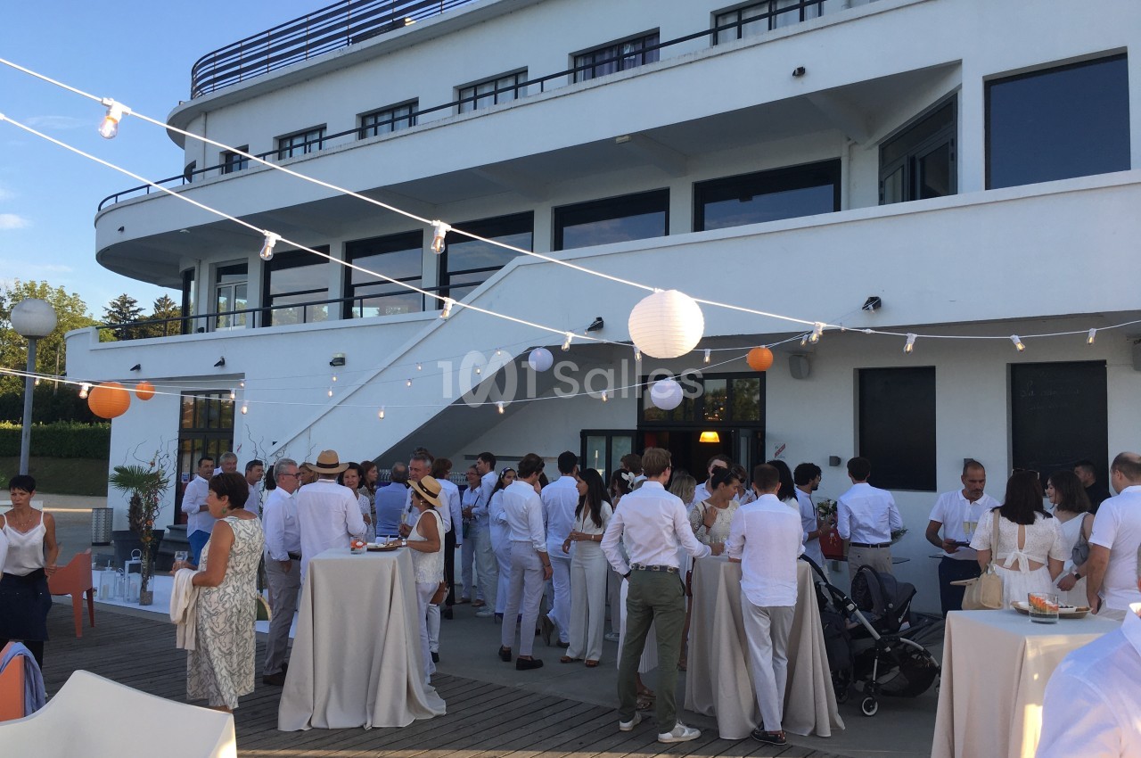 Groupe de personnes en tenue blanche réunies sur une terrasse extérieure, décorée de guirlandes lumineuses et lanternes.