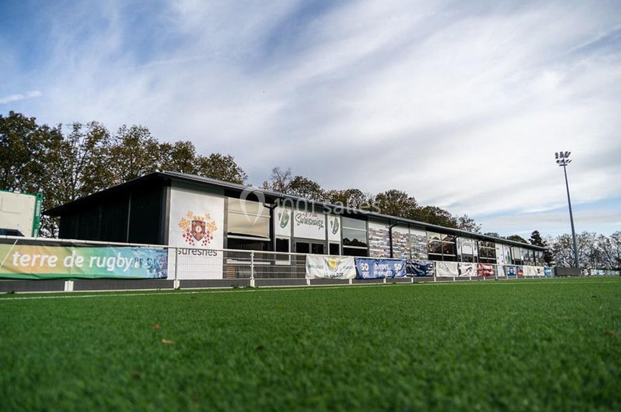 Terrain de rugby avec pelouse synthétique, bordé par un bâtiment et des panneaux publicitaires sous un ciel dégagé.