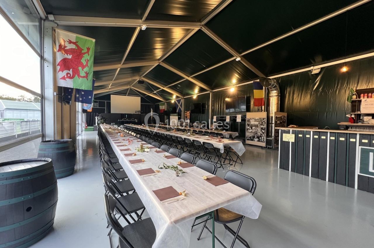 Salle aménagée avec de longues tables dressées pour un événement, décorée de drapeaux et éclairée par des lumières…