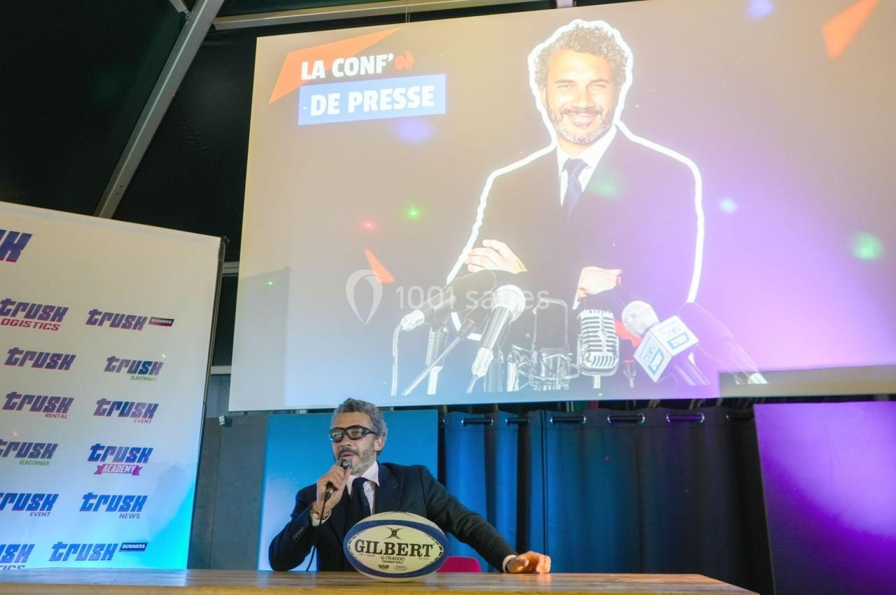 Un homme en costume parle dans un micro lors d'une conférence de presse, avec une projection lumineuse en arrière-plan.