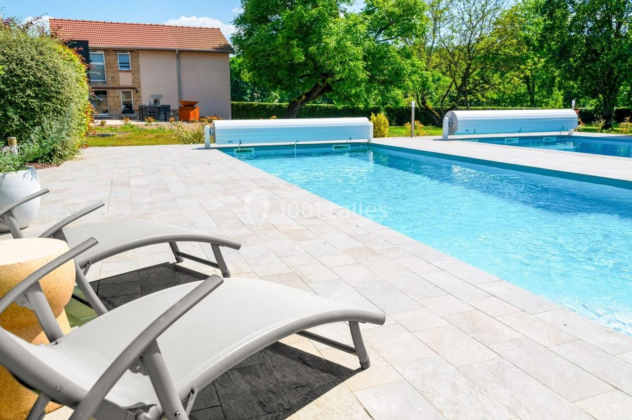 Deux transats gris près d'une piscine entourée de dalles, avec un jardin verdoyant et une maison en arrière-plan.