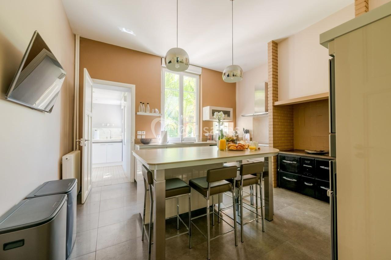 Cuisine moderne avec îlot central, tabourets, électroménagers intégrés, et grande fenêtre donnant sur un jardin.