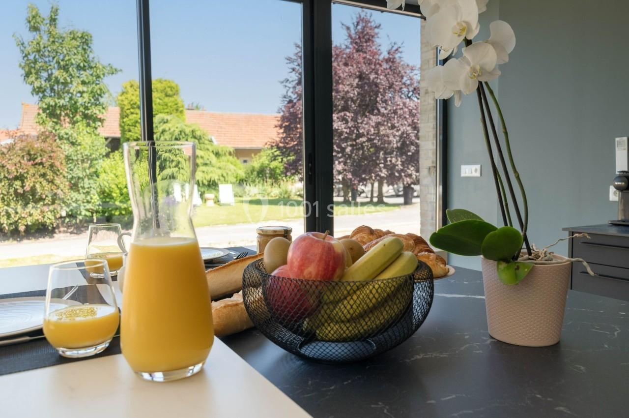 Petit-déjeuner sur une table avec jus d'orange, corbeille de fruits, viennoiseries et une orchidée en arrière-plan lumineux.