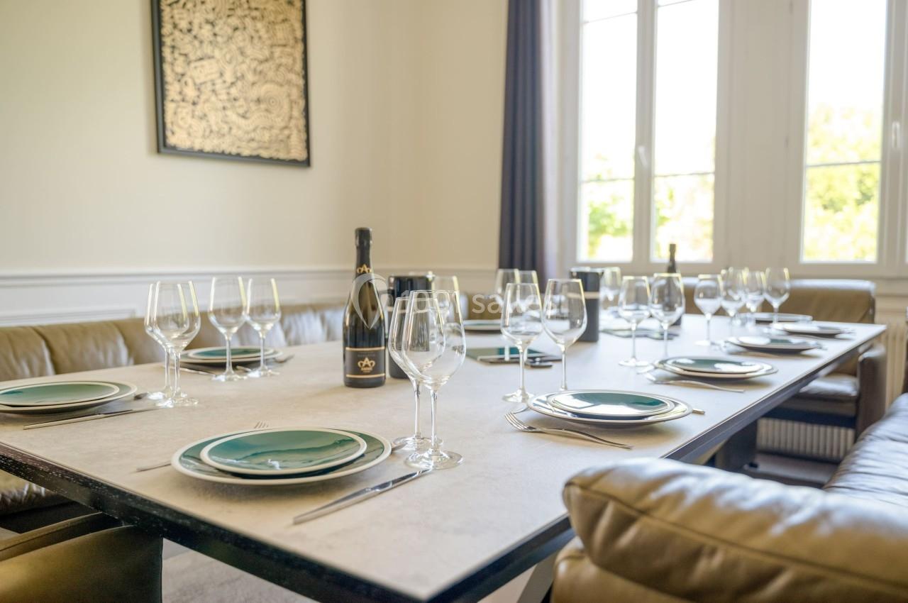 Table dressée avec des assiettes, verres à vin et une bouteille de champagne dans une salle lumineuse avec canapé en cuir.