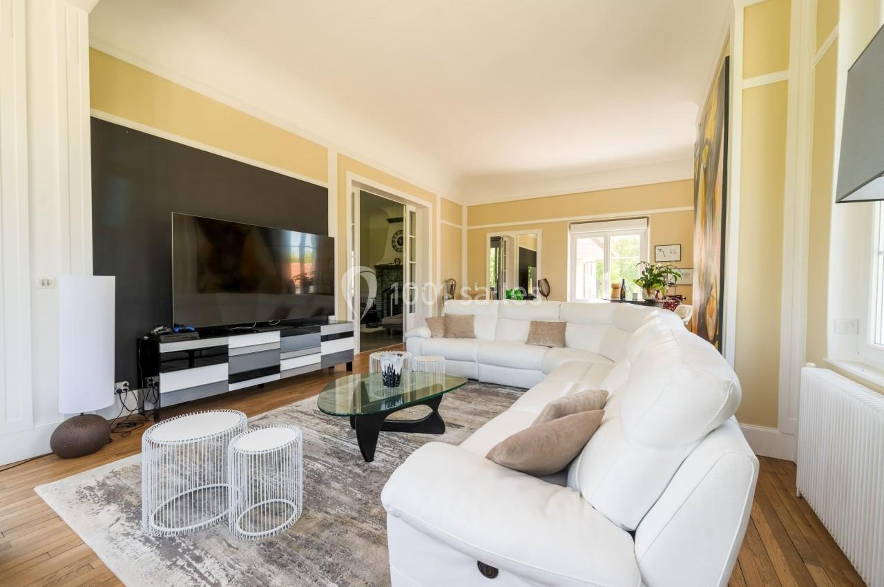 Salon lumineux avec canapé d'angle blanc, table basse en verre, tapis gris et grande télévision sur meuble moderne.