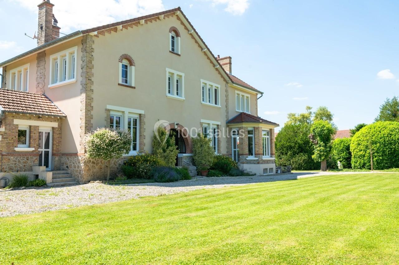 Grande maison en pierre et crépi beige avec jardin verdoyant et allée gravillonnée sous un ciel dégagé.