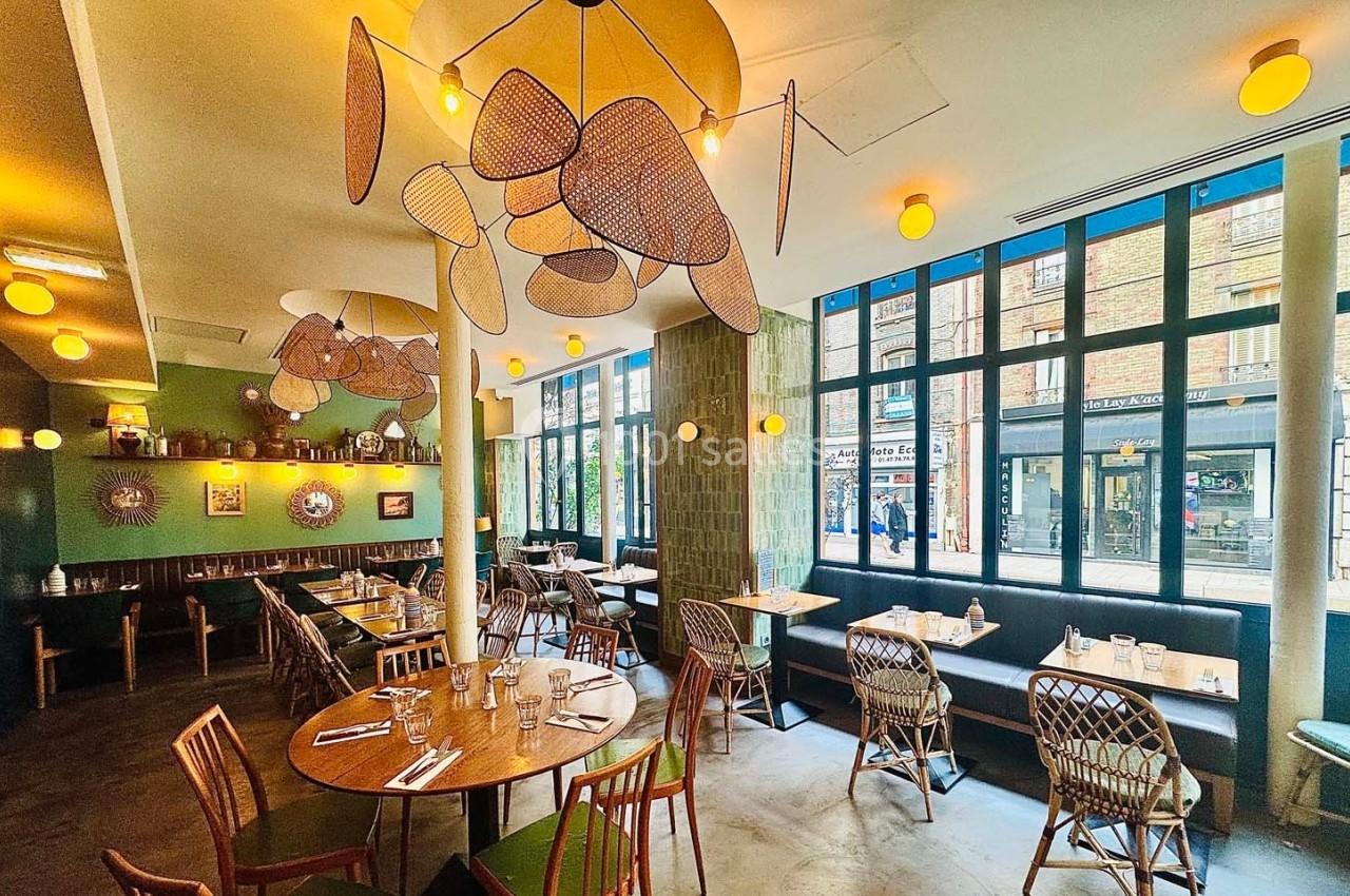 Salle de restaurant lumineuse avec grandes fenêtres, tables en bois, chaises en rotin et décorations suspendues au plafond.