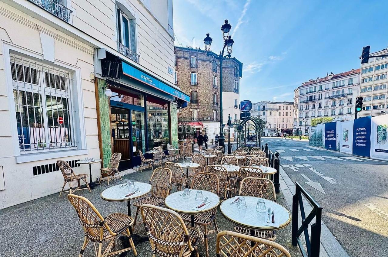 Terrasse de café avec des tables et chaises en osier, située sur un trottoir ensoleillé dans un quartier urbain.