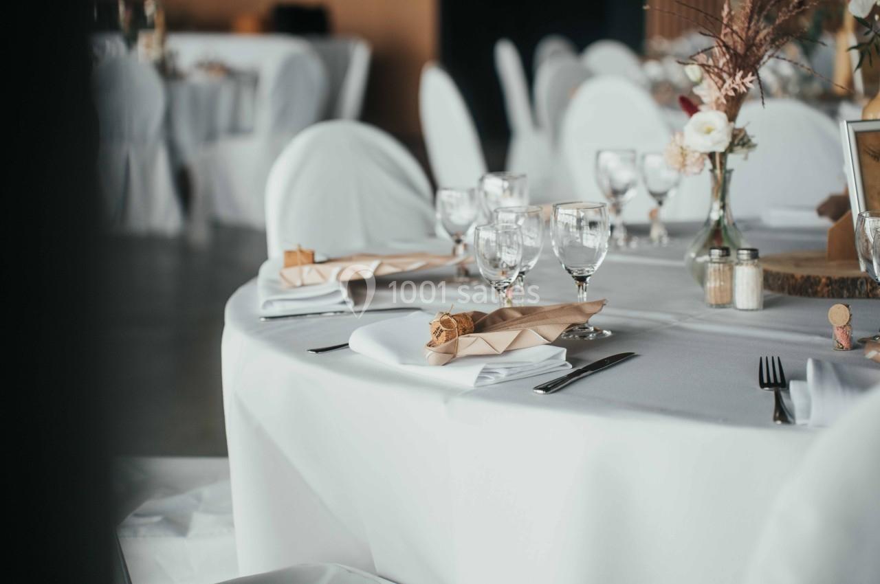 Table dressée avec nappes blanches, serviettes pliées, verres à pied et décorations florales dans une salle de réception.