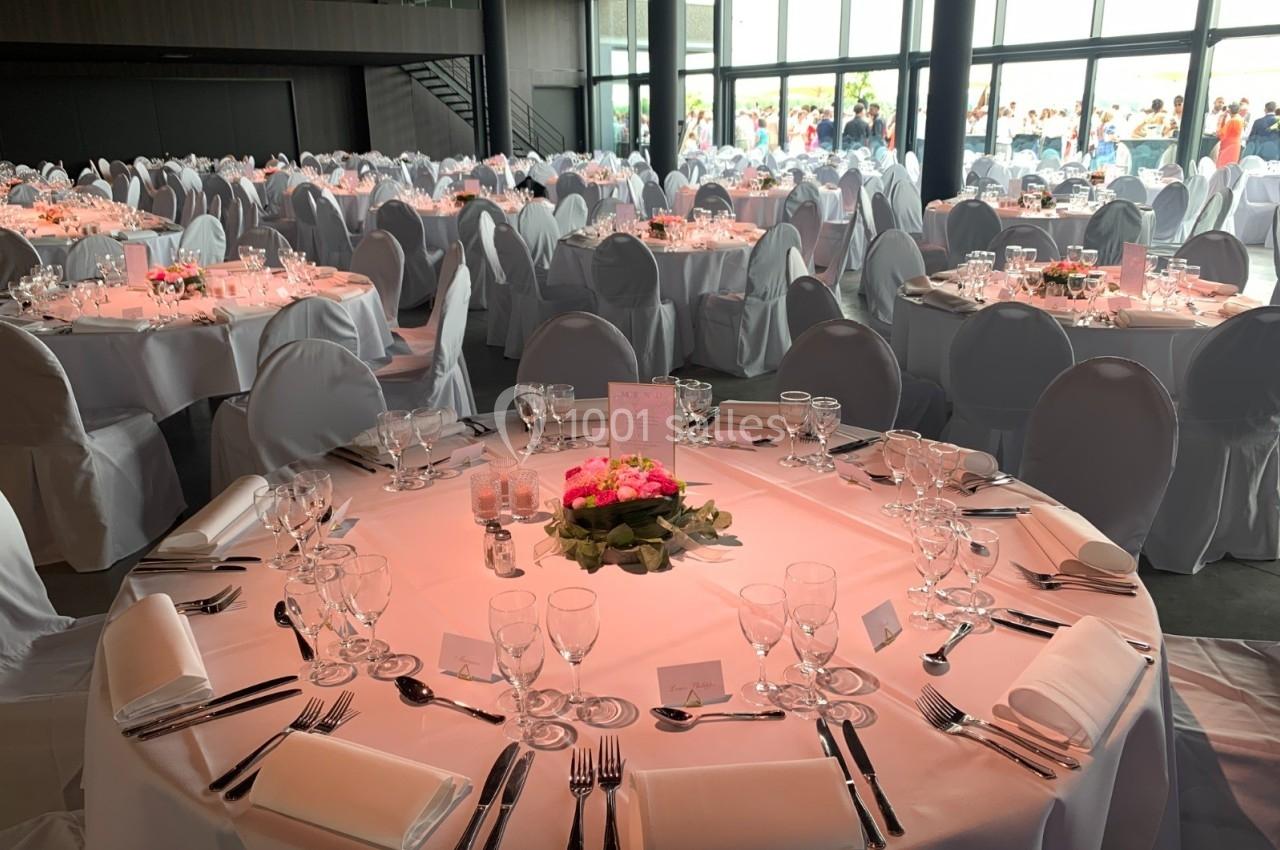 Salle de réception avec tables rondes élégamment dressées, nappes blanches et décoration florale rose.