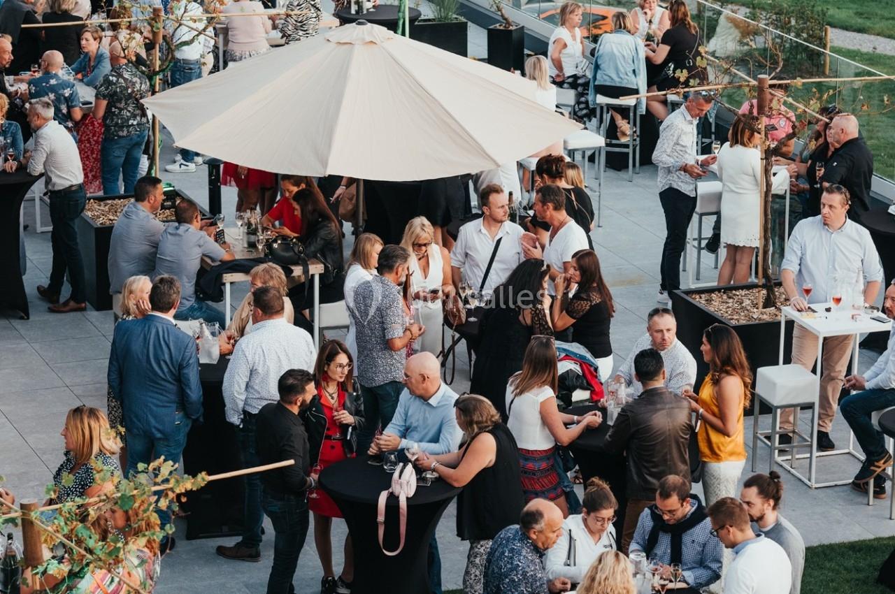 Groupe de personnes rassemblées lors d'un événement en plein air, discutant autour de tables sous un grand parasol.