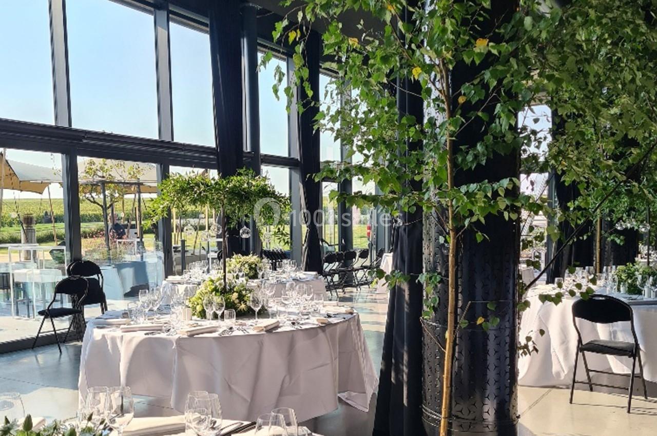 Salle lumineuse avec grandes baies vitrées, tables rondes dressées pour un repas et décoration végétale.