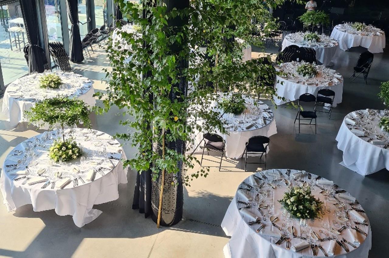 Salle de réception lumineuse avec des tables rondes décorées de nappes blanches, couverts et arrangements floraux.