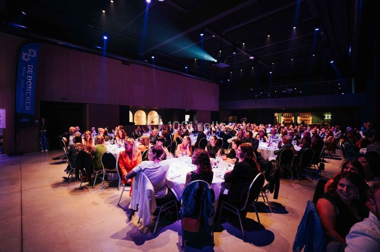 Salle de réception avec des invités assis à des tables rondes, éclairée par des lumières tamisées.