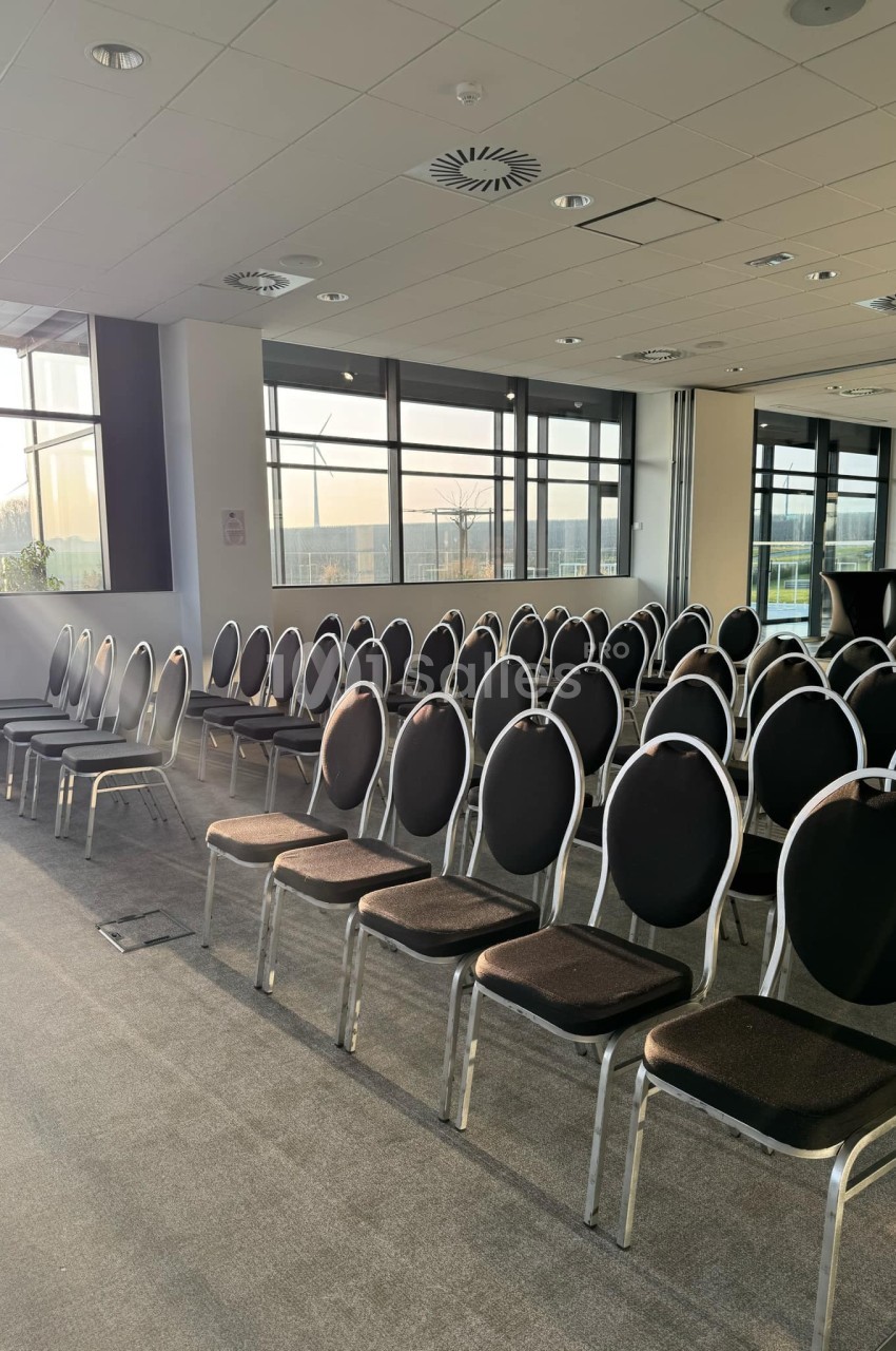 Salle de conférence vide avec des rangées de chaises noires alignées, éclairée par la lumière naturelle d'une grande fenêtre.