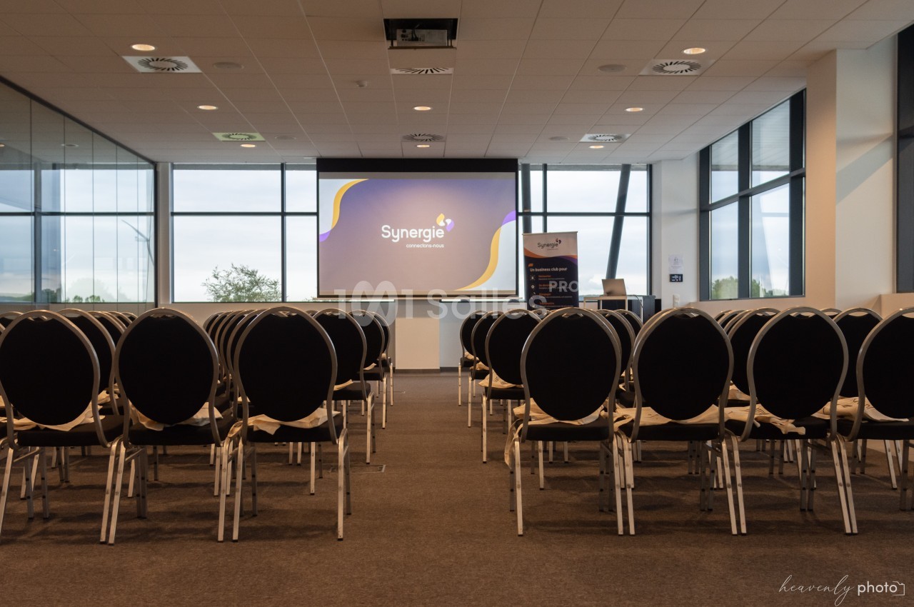 Salle de conférence vide avec des rangées de chaises noires face à un écran de projection allumé.