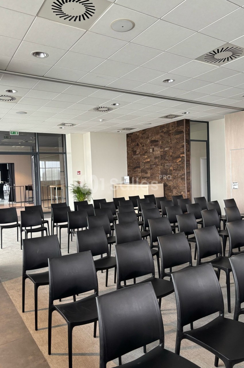 Salle de conférence moderne avec rangées de chaises noires alignées, murs clairs et comptoir en arrière-plan.
