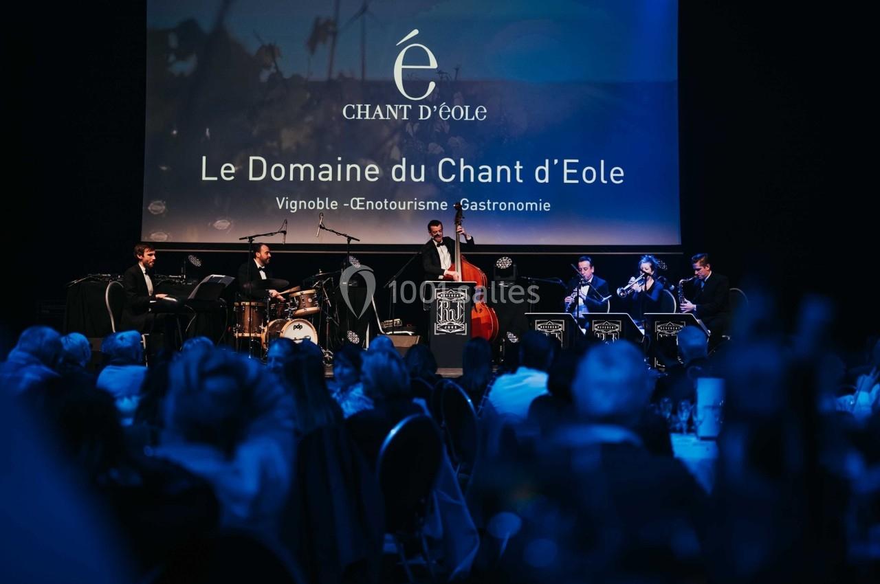 Un groupe de musiciens joue sur scène lors d'un événement dans une salle sombre avec un écran projetant un logo.