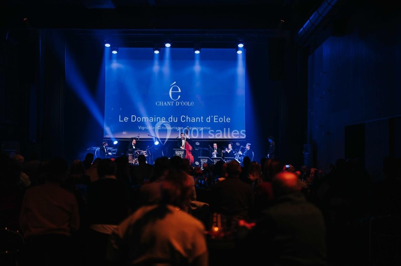 Concert en intérieur avec un groupe sur scène, éclairé par des projecteurs, devant un public assis.