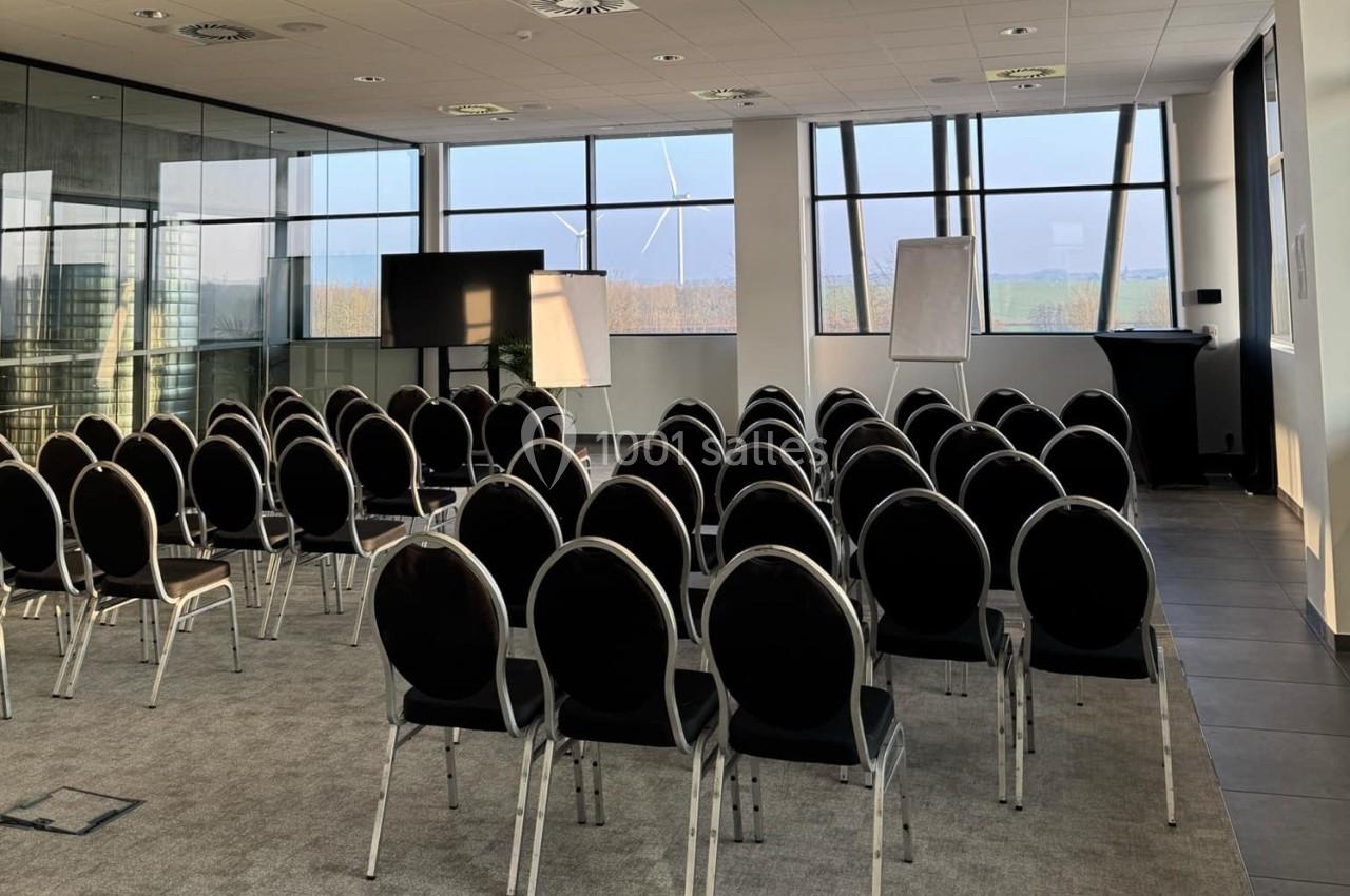 Salle de conférence lumineuse avec des rangées de chaises noires, un écran, des tableaux blancs et de grandes fenêtres.
