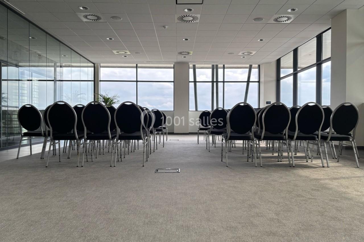 Salle de conférence lumineuse avec des rangées de chaises noires alignées face à une grande baie vitrée.