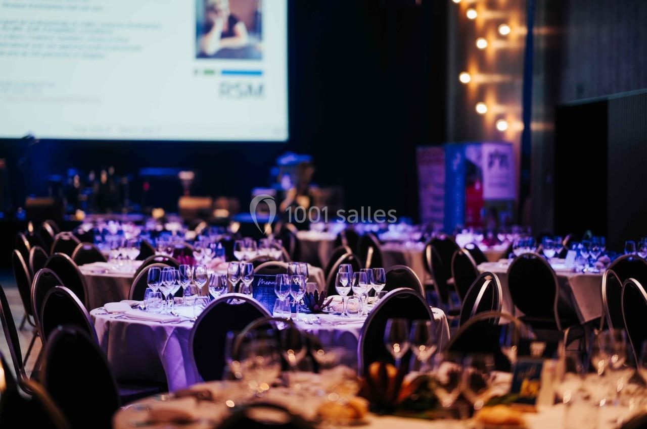 Salle de réception avec tables rondes dressées, verres et couverts, éclairage tamisé et écran en arrière-plan.