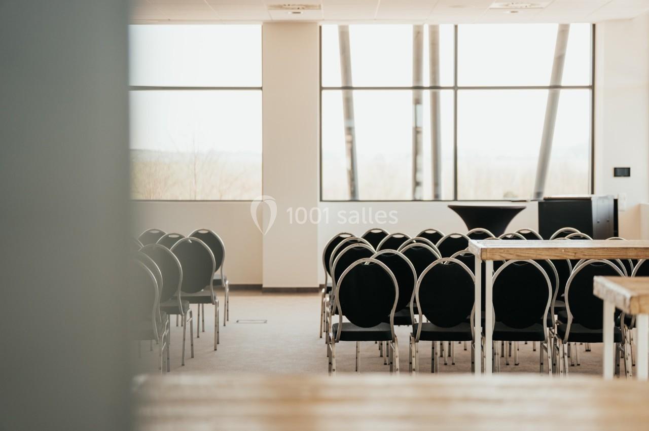 Salle de conférence lumineuse avec des rangées de chaises noires et des tables en bois, vue sur de grandes fenêtres.