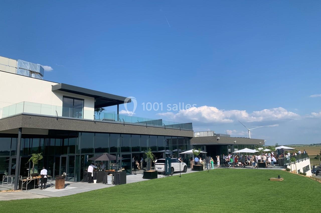 Terrasse moderne avec tables et parasols, entourée de pelouse, sous un ciel bleu dégagé.