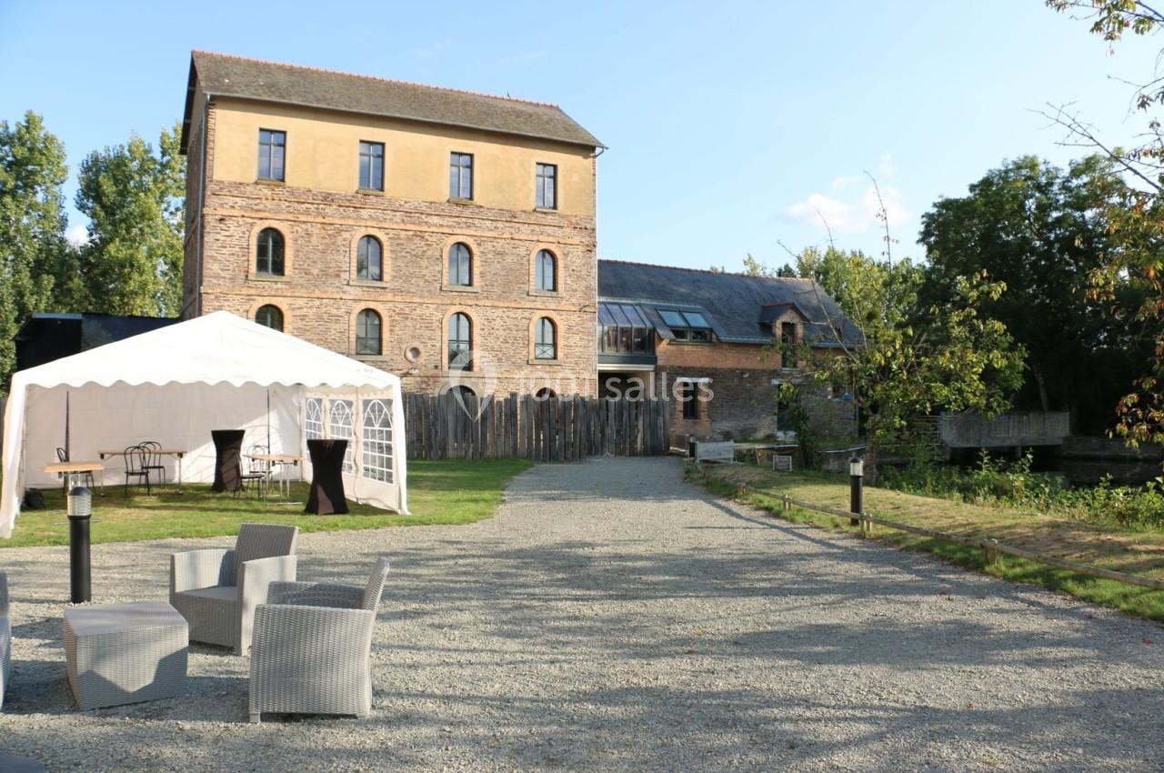 Bâtiment en briques avec une tente blanche et des fauteuils sur une allée gravillonnée, entouré de verdure.