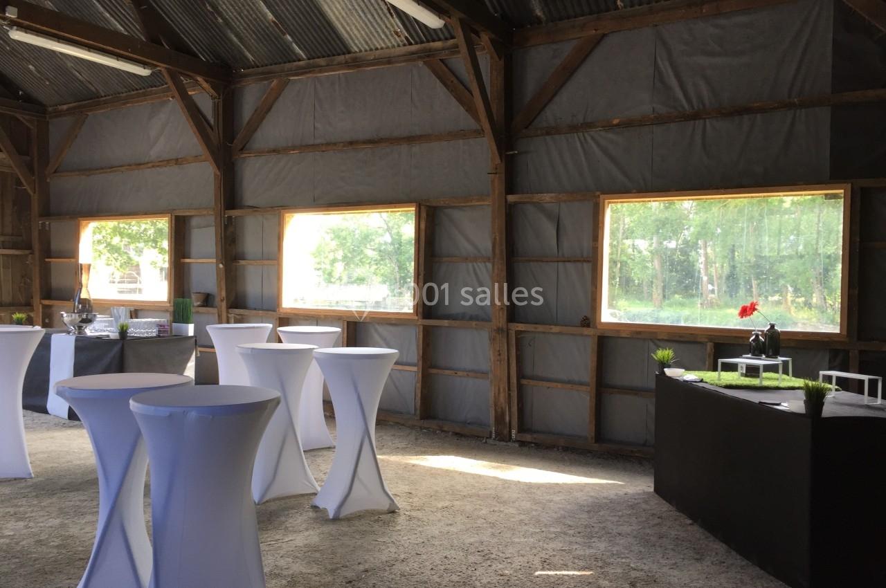 Intérieur d'une grange aménagée avec des tables hautes drapées de blanc et des fenêtres donnant sur un paysage verdoyant.