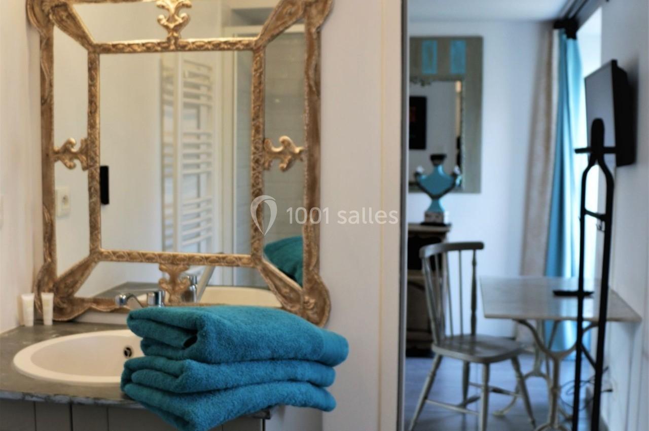 Salle de bain avec un lavabo, un miroir doré orné, des serviettes bleues pliées et vue sur une pièce adjacente.