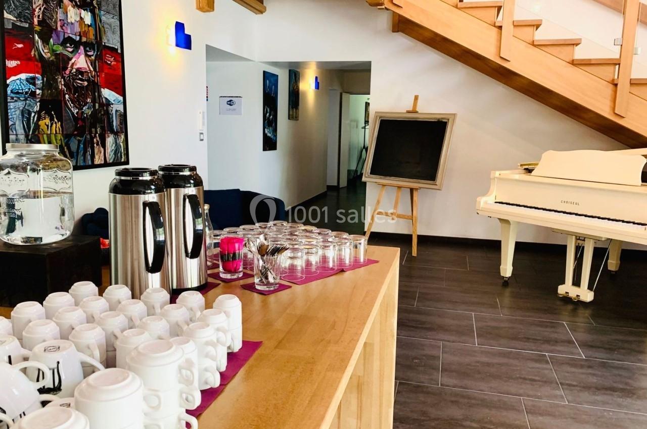Buffet avec tasses, thermos et verres sur une table en bois, dans une pièce lumineuse avec escalier et piano blanc.
