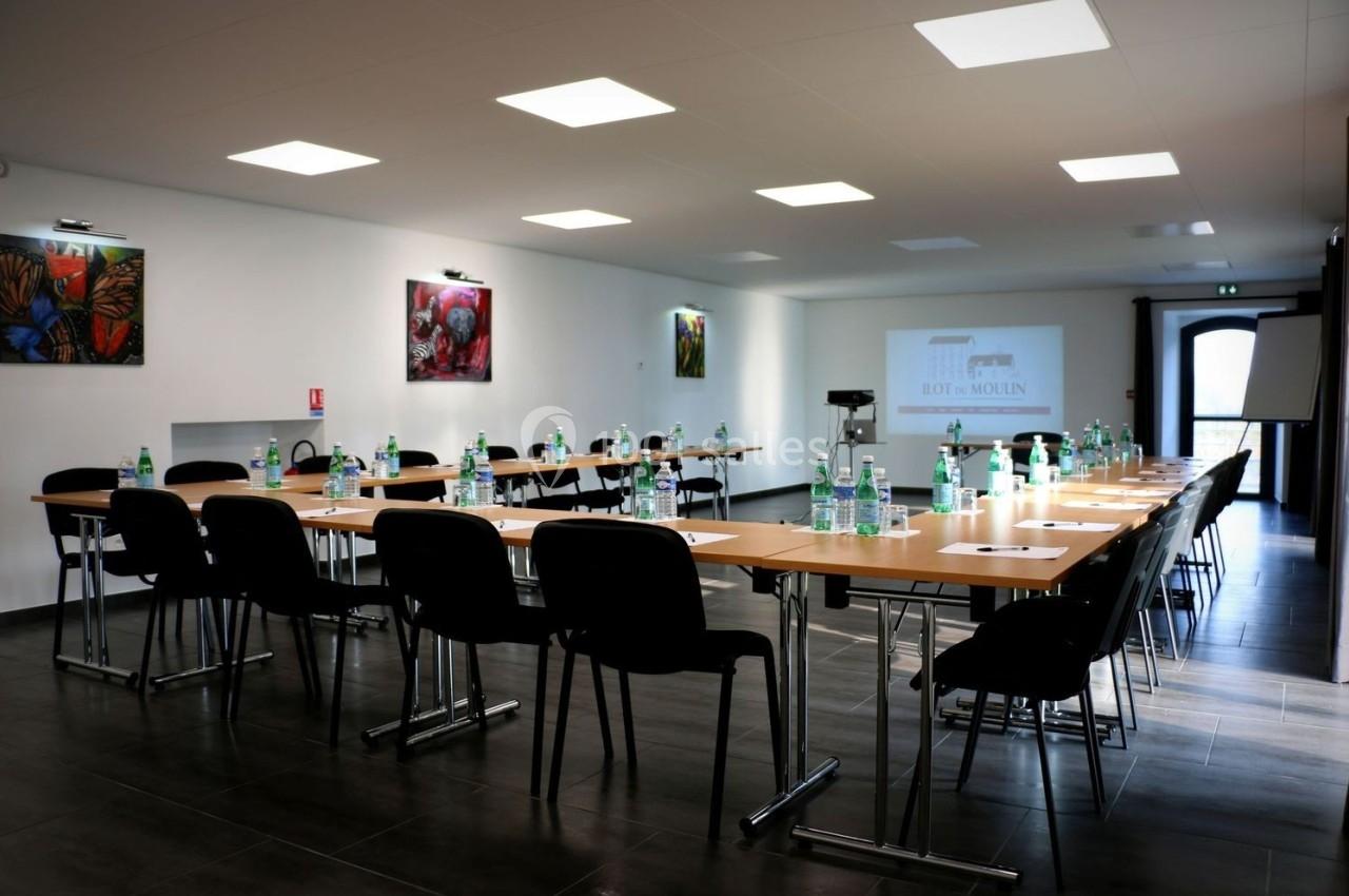 Salle de réunion lumineuse avec tables disposées en U, chaises noires, bouteilles d'eau et documents sur les tables.