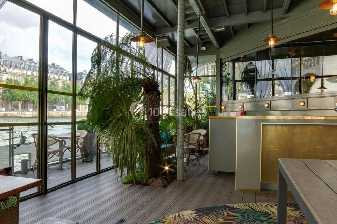 Intérieur d'un espace lumineux avec baies vitrées donnant sur une rivière, décoré de plantes et de mobilier en bois.