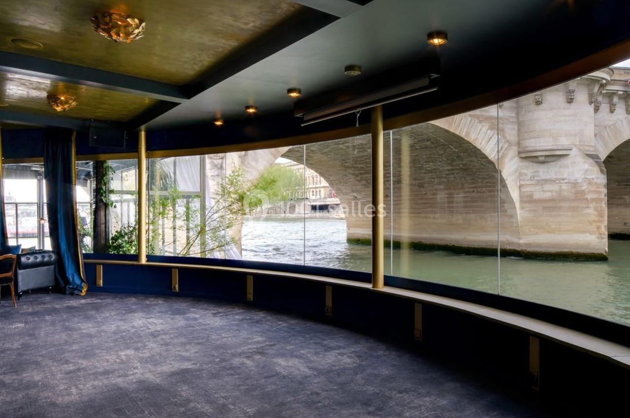 Vue intérieure d'une salle avec de grandes baies vitrées donnant sur un pont en pierre et une rivière.